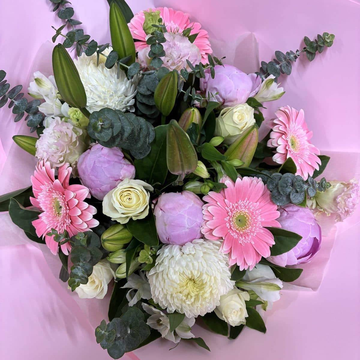 Pastel bouquet in blush pink wrap: pink gerberas, lilac peonies, white chrysanthemums, cream roses and eucalyptus, photographed from above