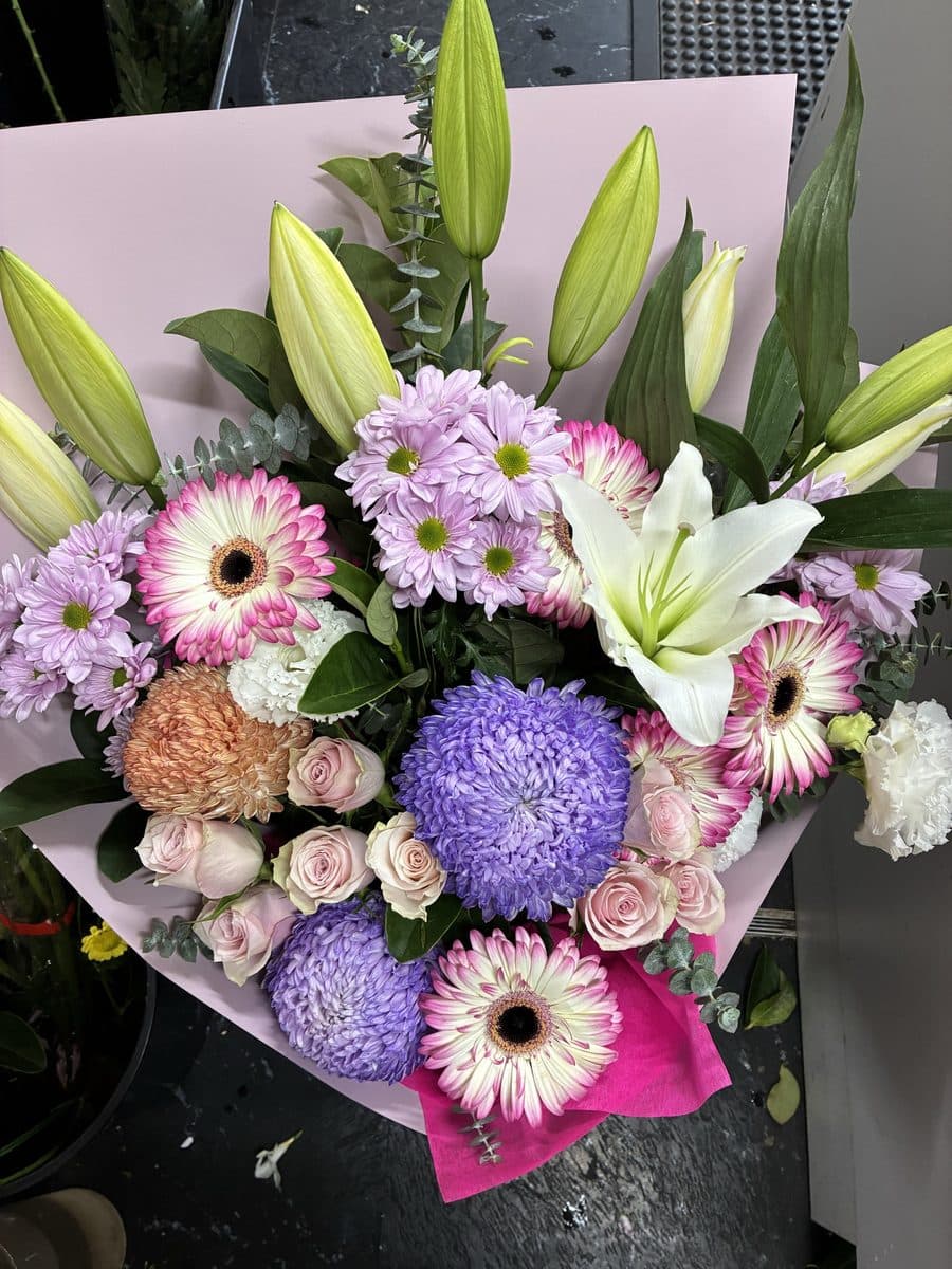 Bouquet in pink wrap with white oriental lilies, pale pink gerberas, a purple chrysanthemum, lilac daisies and spray roses