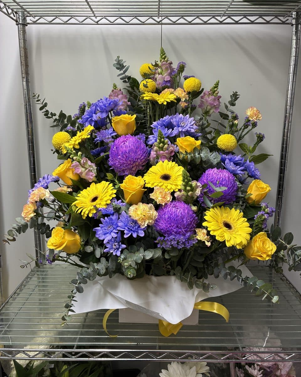 Large box arrangement of purple chrysanthemums, yellow gerberas, yellow roses, snapdragons and eucalyptus with a yellow ribbon