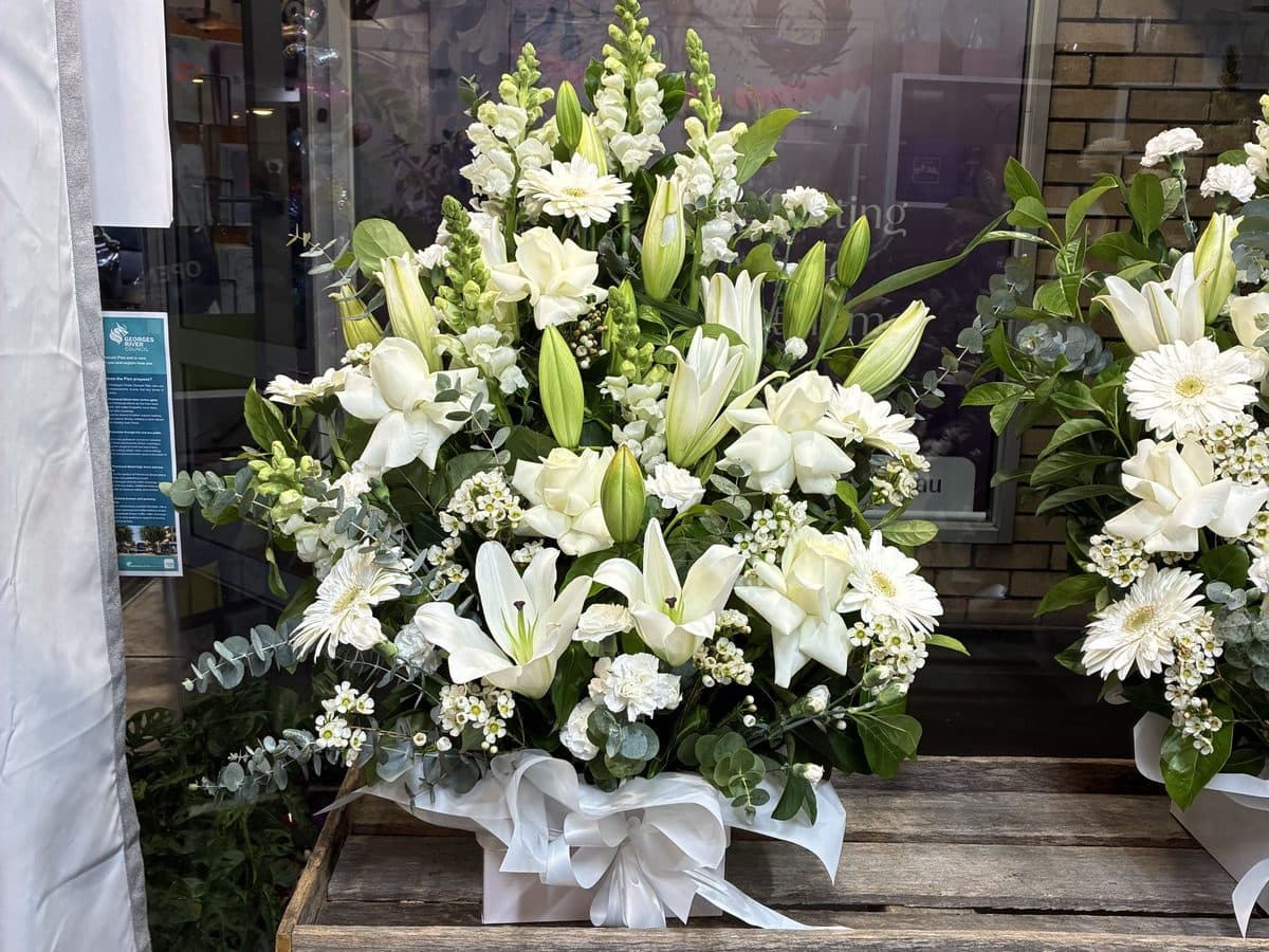 Two tall white arrangements of oriental lilies, chrysanthemums and snapdragons on a timber bench in the shop window
